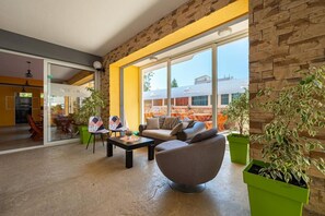 Un área de descanso moderna con un sofá, sillones y una mesa de centro. Hay plantas en macetas y un separador de vidrio que divide el área de descanso de un comedor.