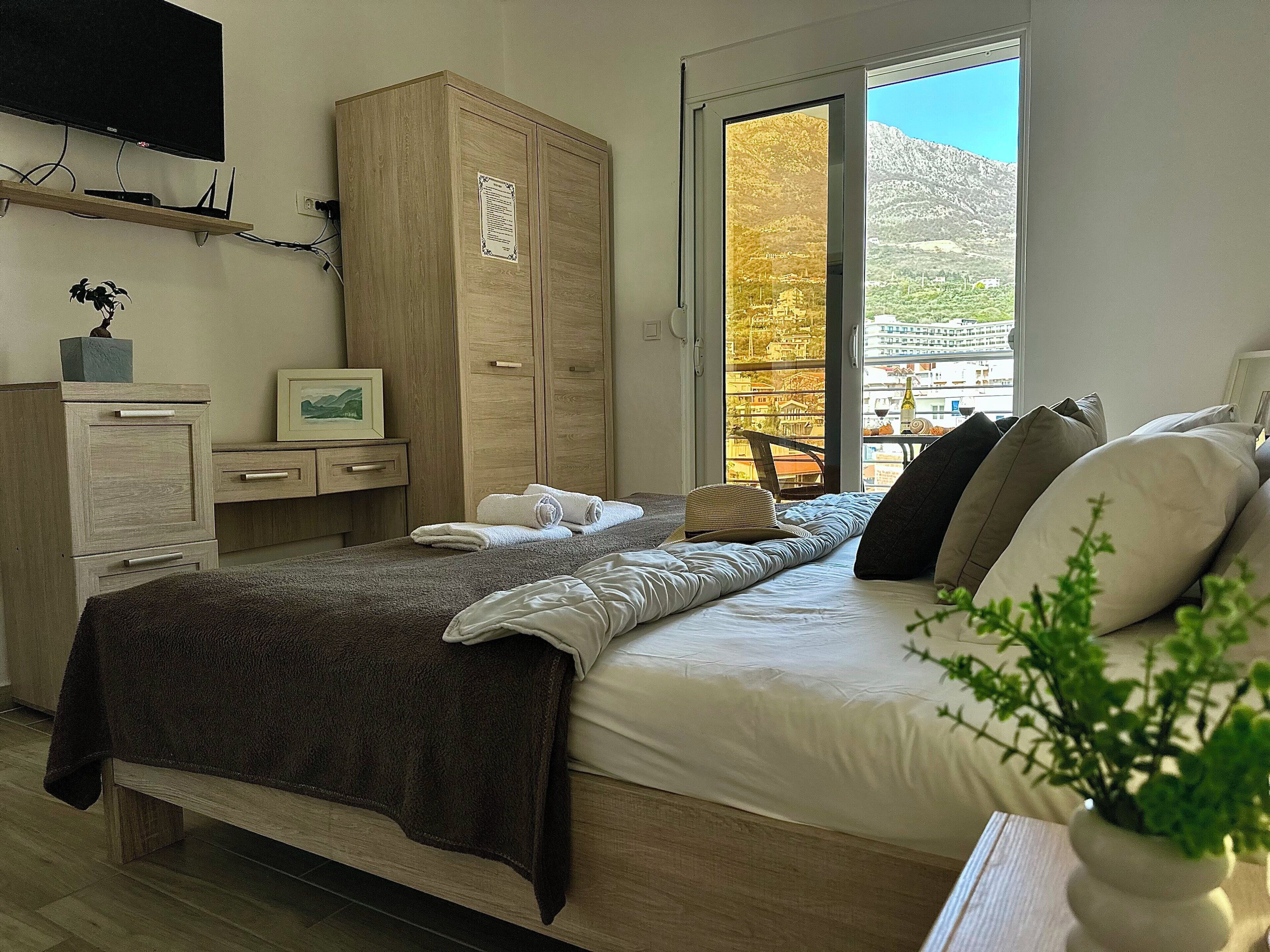 Quarto com cama, guarda-roupa, escrivaninha, televisão e janela com vista para a montanha.