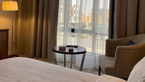 A hotel room with a bed, a desk, a chair, a lamp, and a view of the street through sheer curtains. - Shorfa for Hotel Apartments Al-Shorfa (Riyadh)