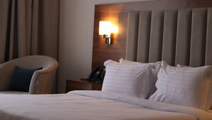 A hotel room with a bed, a chair, a nightstand with a phone, and a wall-mounted lamp. - Shorfa for Hotel Apartments Al-Shorfa (Riyadh)