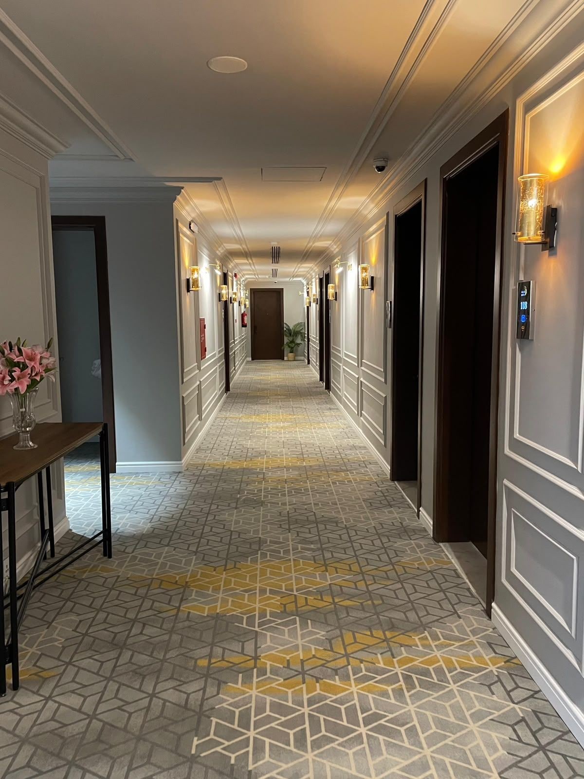 A hotel corridor with a patterned floor, doors on both sides, and wall-mounted light fixtures.