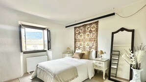 A bedroom with a large bed, a nightstand, a dresser, a mirror, and a window with a view of buildings. - Casa Medieval en el Centro de Montblanc (Montblanc)