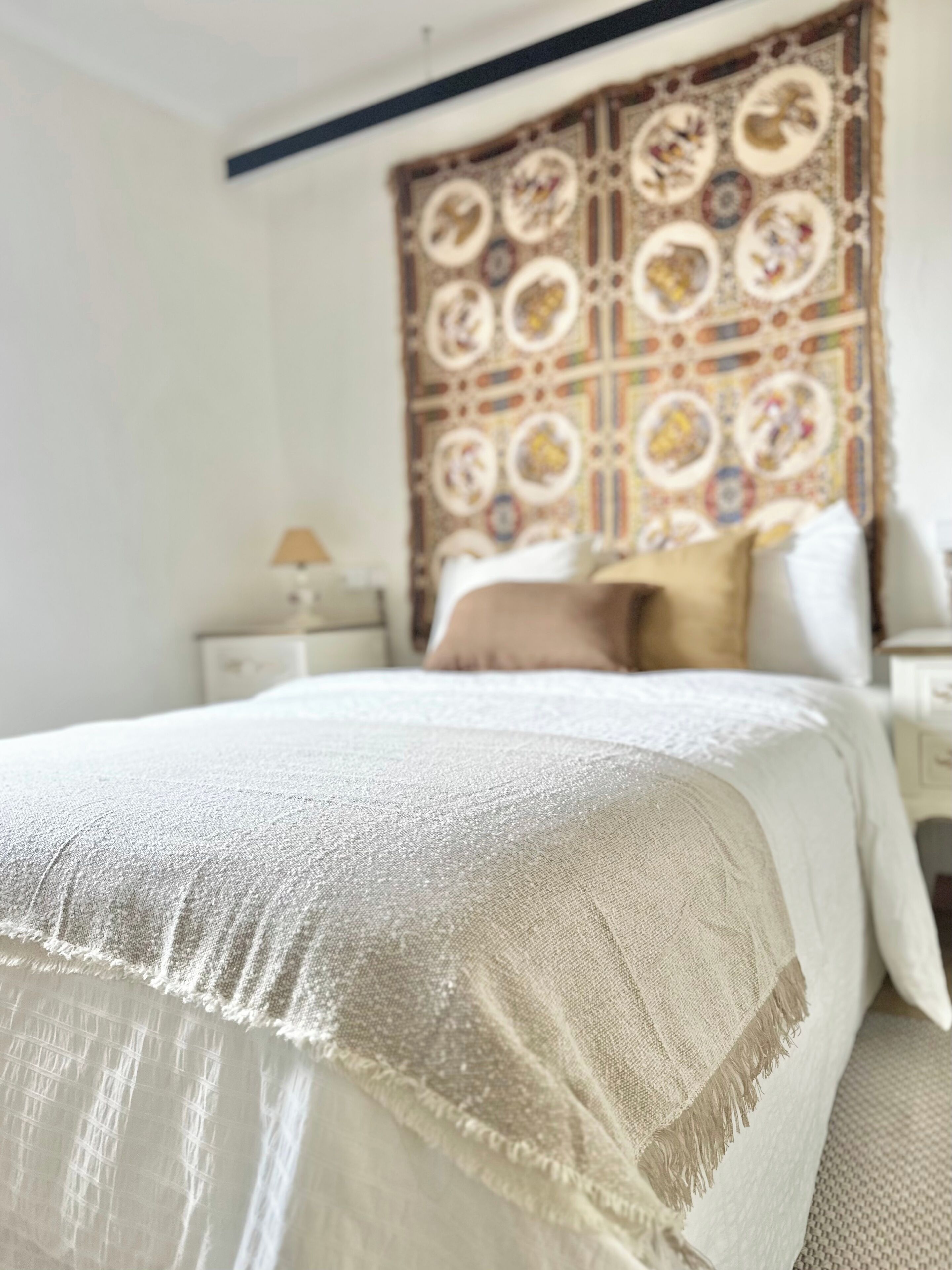 Una cama bien hecha con colcha blanca y color crema, un tapiz decorativo en la pared y una mesita de noche con lámpara.