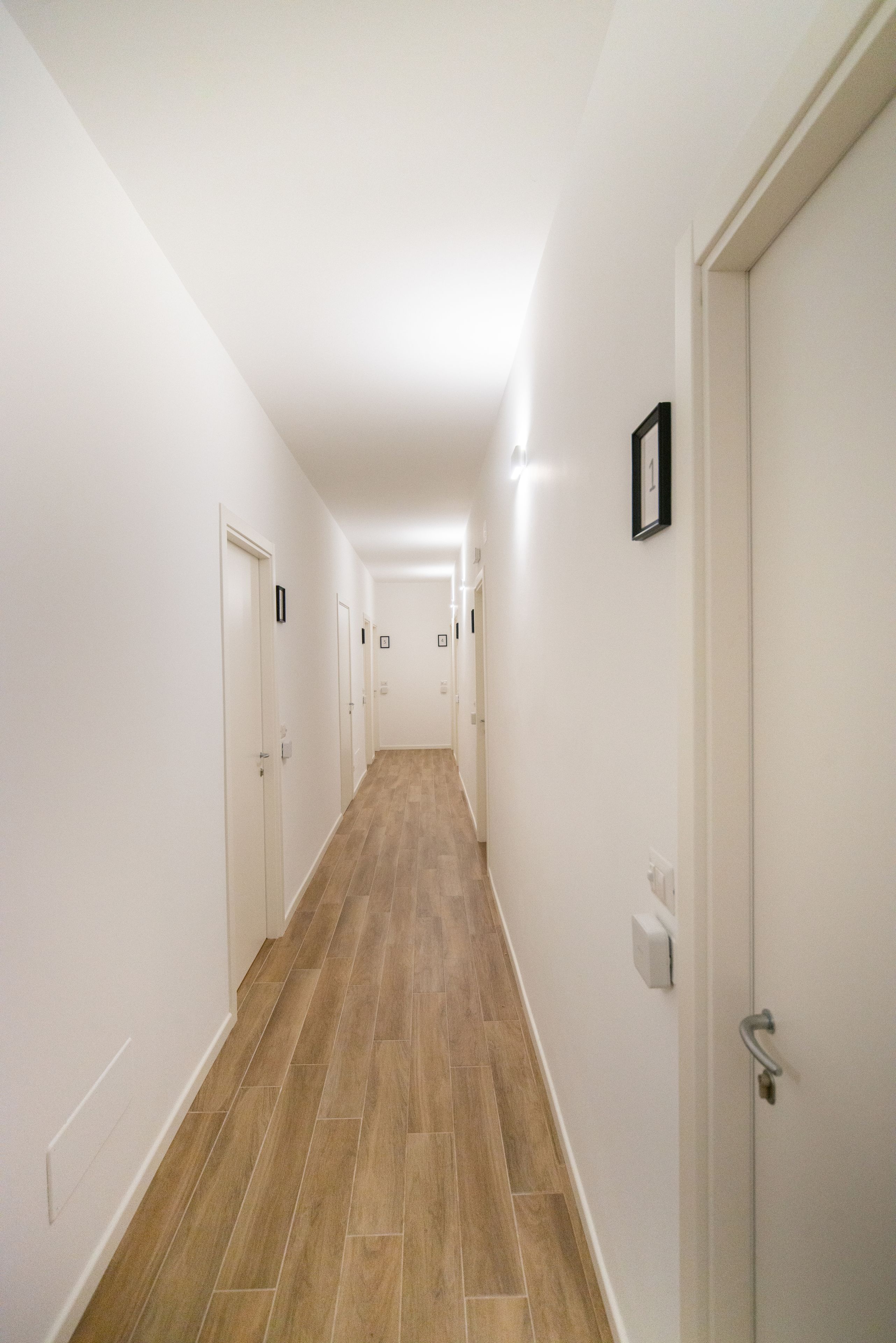 A long, narrow hallway with white walls and wooden flooring, leading to several doors.