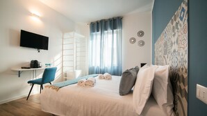 A hotel room with a bed, a desk with a chair, a TV mounted on the wall, and a window with curtains.