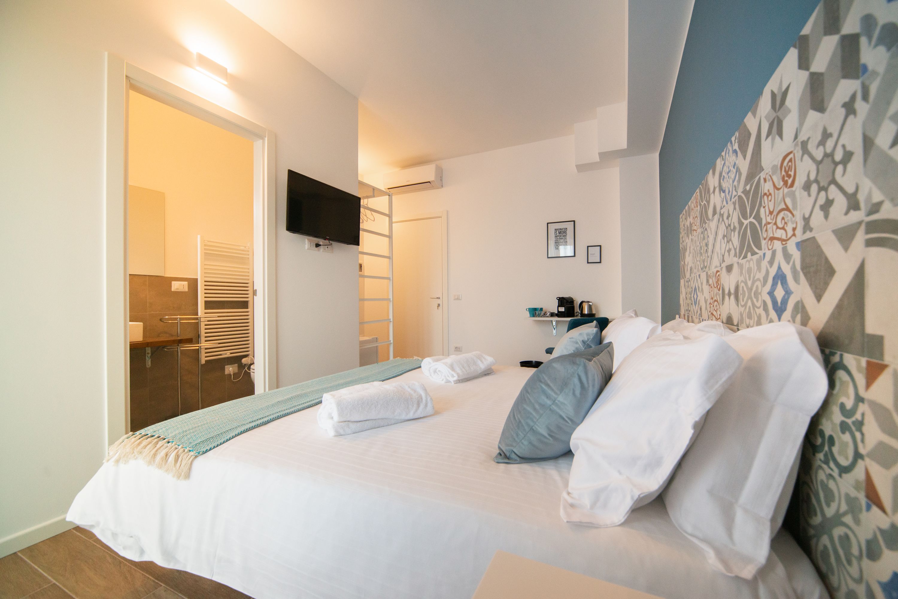 A neatly made bed with white linens and a blue throw blanket, a flat-screen TV mounted on the wall, and a patterned wall design.