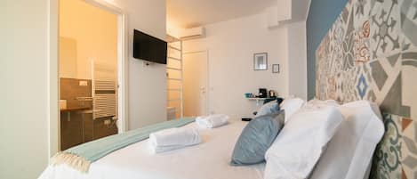 Una cama bien hecha con ropa de cama blanca y una manta azul, un televisor de pantalla plana montado en la pared y un diseño de pared estampado.
