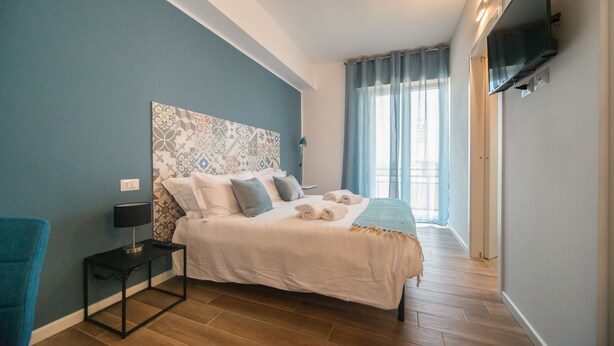 A bedroom with a bed, a nightstand, a TV mounted on the wall, and a window with curtains. - Casa Stazione (Verona)