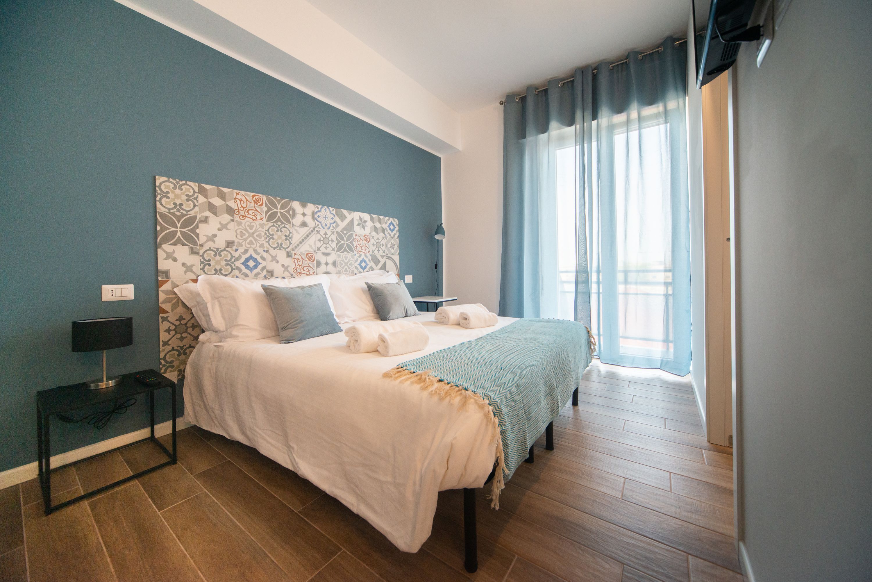 Un dormitorio con una cama grande, una mesita de noche, una lámpara y una ventana con cortinas.