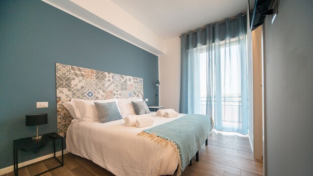A bedroom with a large bed, a bedside table, a lamp, and a window with curtains.