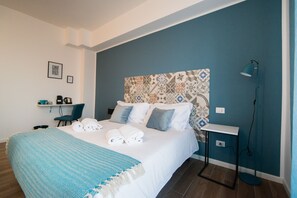 A modern hotel room with a neatly made bed, a desk with a chair, a standing lamp, and a wall with patterned tiles. - Casa Stazione (Verona)