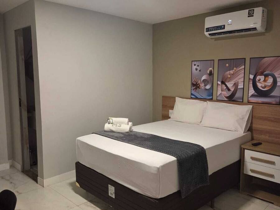 A modern hotel room with a bed, a nightstand, an air conditioning unit, and artwork on the wall.