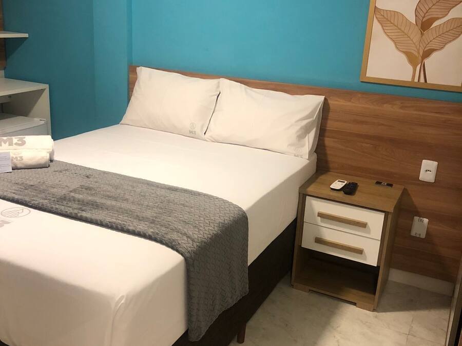Una cama bien tendida con una manta gris, una mesita de noche de madera con un teléfono y una lámpara pequeña, y una pared azul con una obra de arte con diseño de hojas.