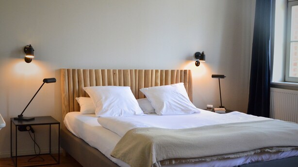 A neatly made bed with a wooden headboard, two bedside tables with lamps, and a window with curtains. - Double room in the Gasthaus am Finowkanal (former school from 1875). (Wandlitz)