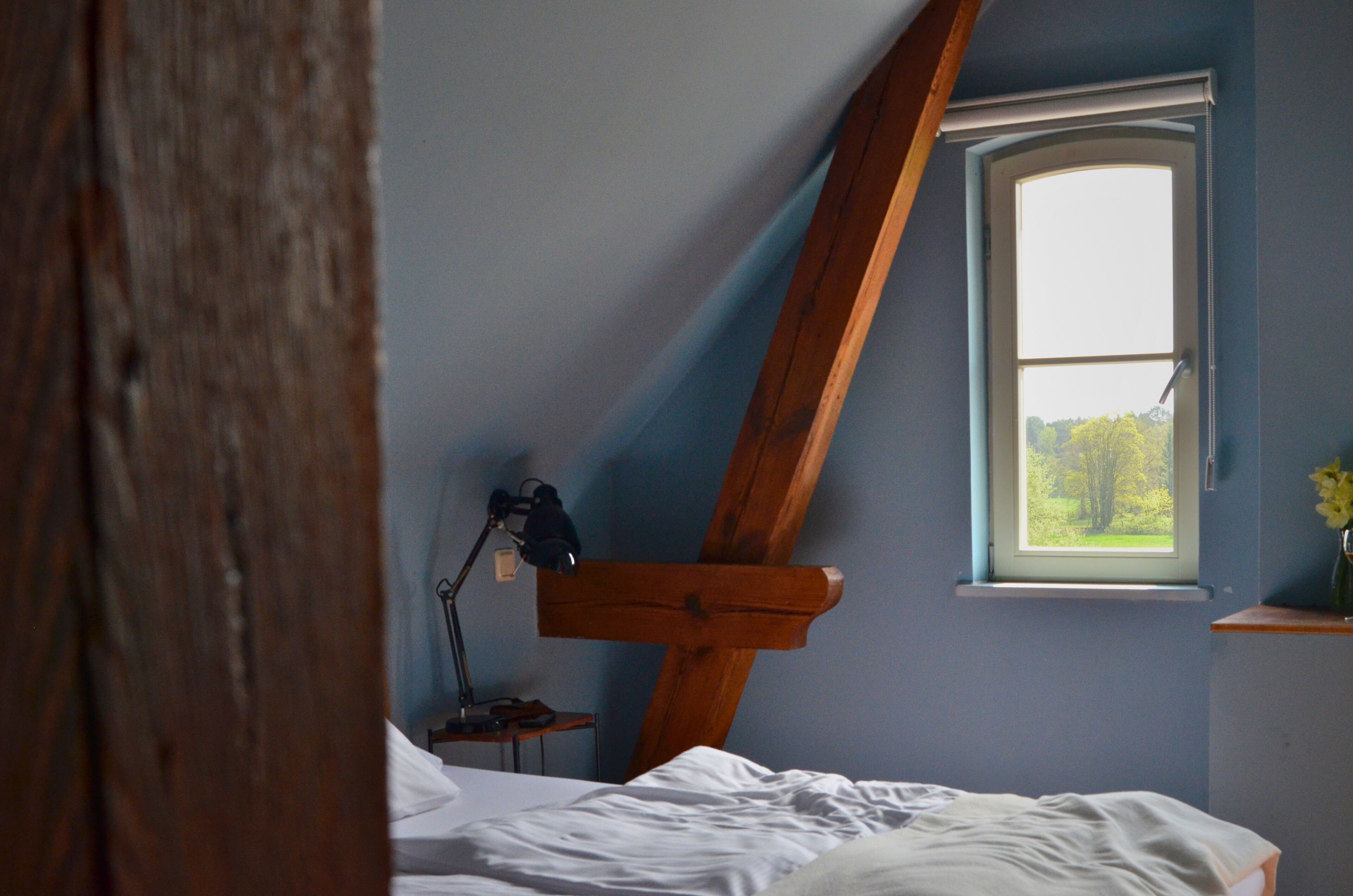A bedroom with a sloped ceiling, a window with a view of greenery, a bedside table with a lamp, and a wooden beam structure.