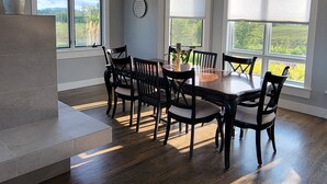 Een eethoek met een houten tafel en stoelen, grote ramen met zonwering en een decoratieve plafondlamp.