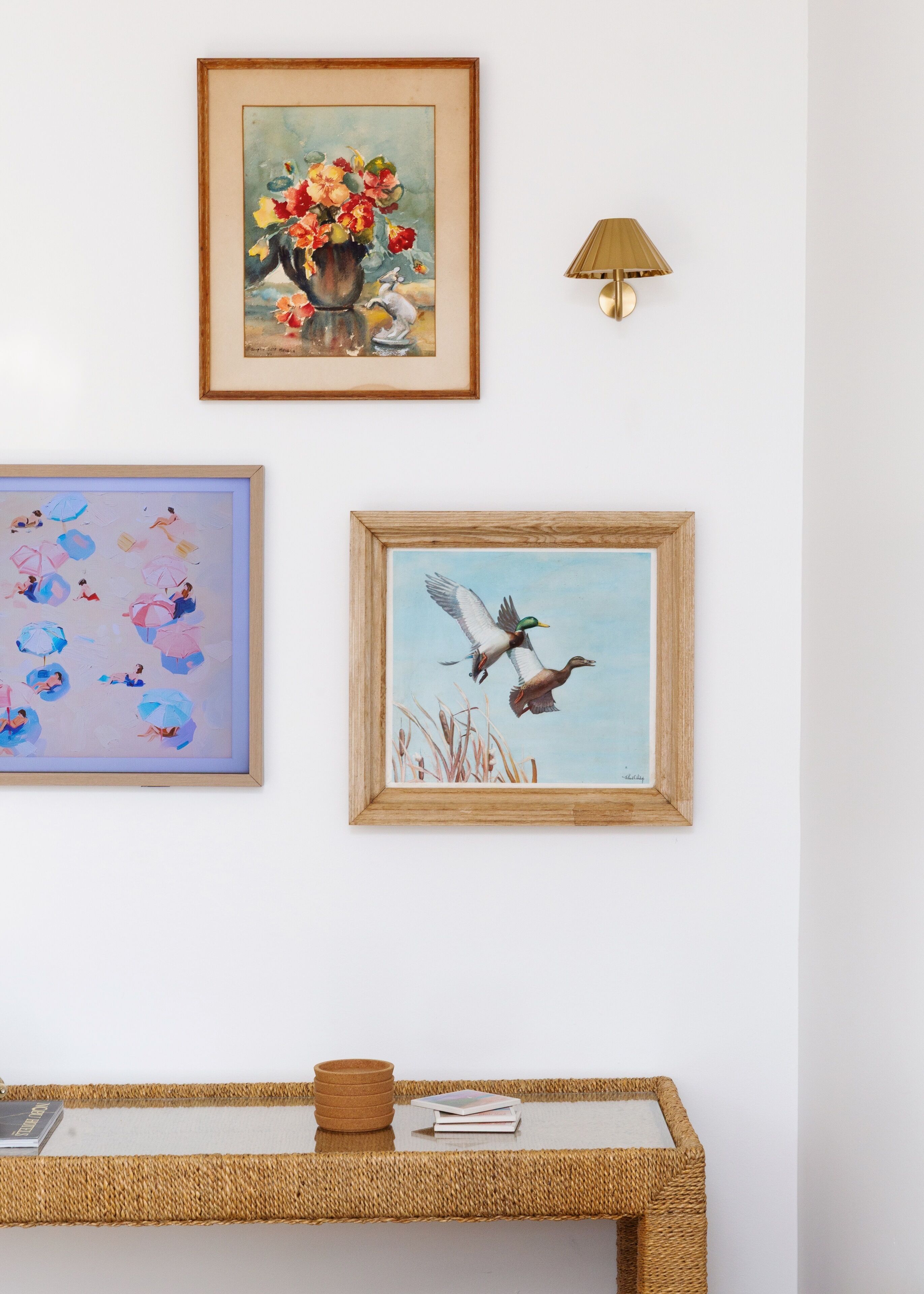 Een muur met drie ingelijste kunstwerken, een lamp en een geweven bijzettafeltje met een boek en een schaal.