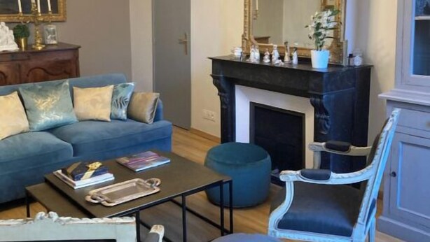 A living room with a blue sofa, a coffee table, a blue ottoman, a white armchair, a fireplace, and a large mirror. - Your cosy, quiet suite in the heart of Carcassonne! (Carcassonne)