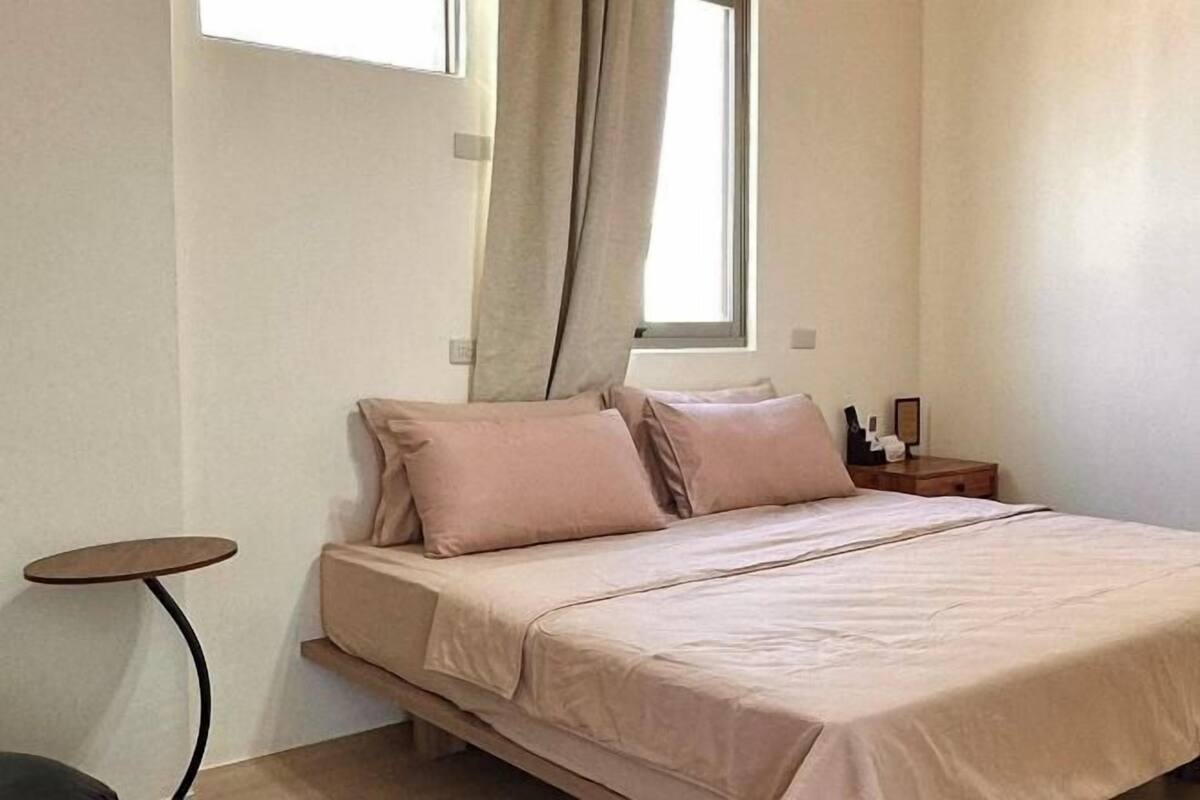 Un dormitorio con una cama, una mesita redonda pequeña, una mesita de noche y una ventana con cortinas.