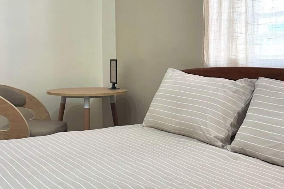 Una cama con ropa de cama a rayas, una mesita redonda pequeña y una silla en una habitación con paredes lisas.