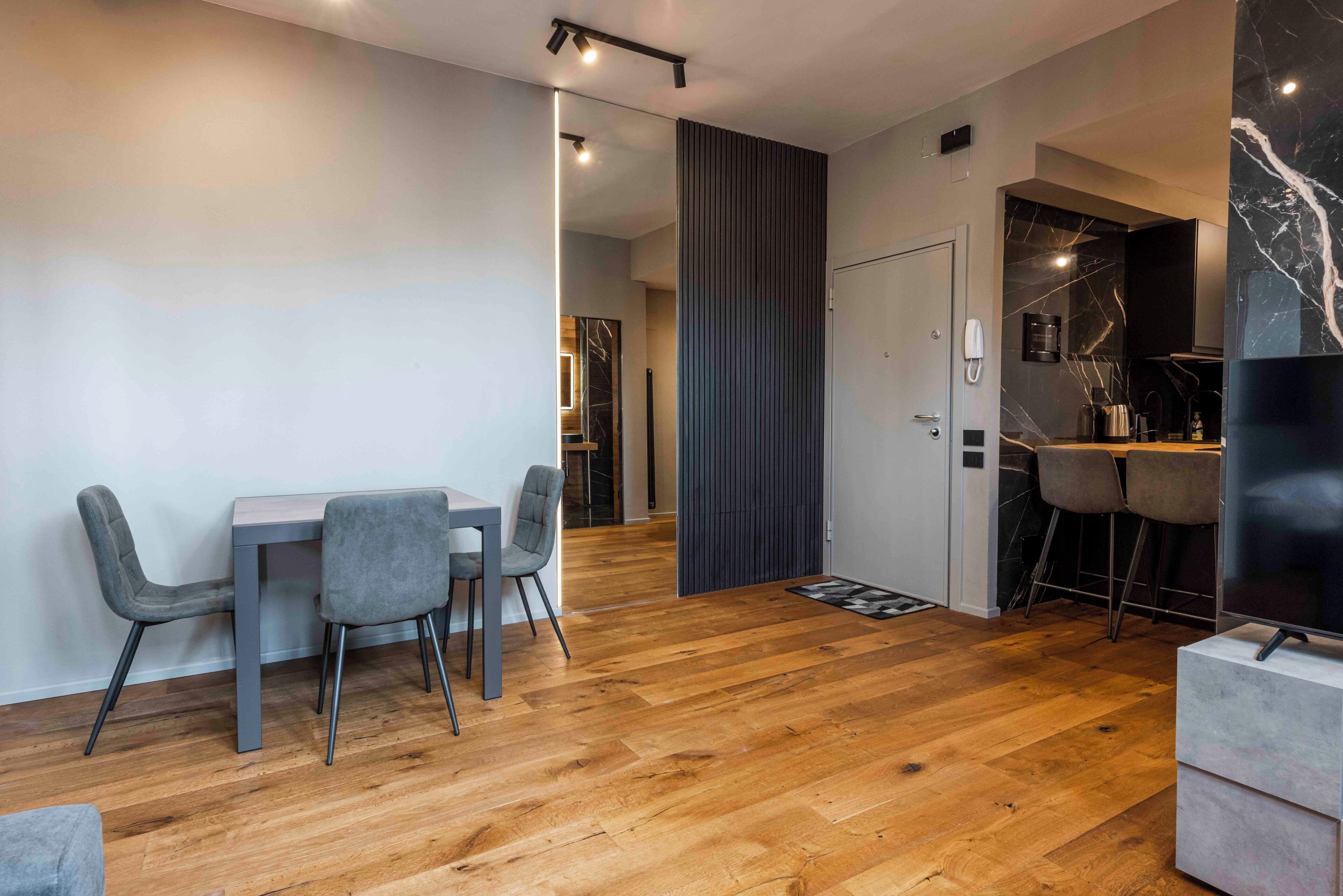 A modern living space with a wooden floor, a dining area with two chairs, a kitchen with a black countertop, and a large TV.