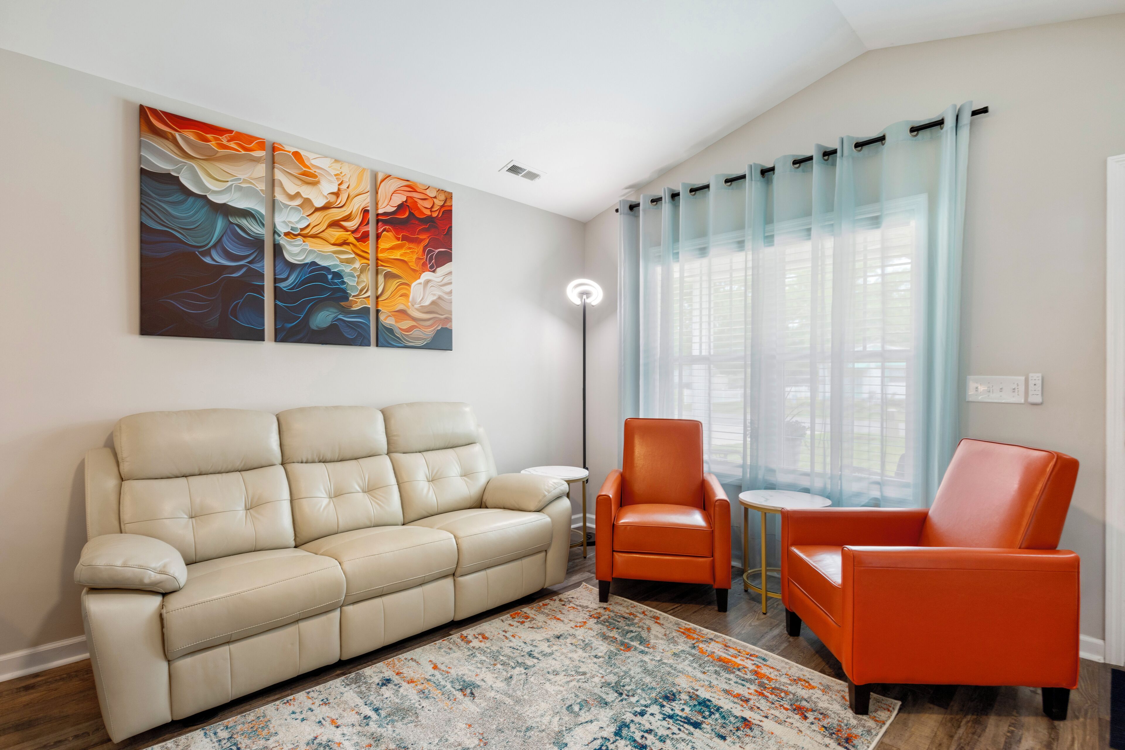 A living room with a beige sofa, two orange armchairs, a coffee table, a floor lamp, and abstract art on the wall.