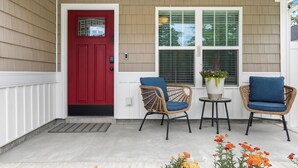 A porch with a red door, two wicker chairs, a small table, and a potted plant with flowers. - Contemporary and Relaxing Getaway - 10 minute walk to the beach (Oak Island)