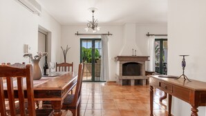 A dining area with a wooden table, chairs, a vase with flowers, a fireplace, and a window with curtains. - Villa 'Anogia  1' with Mountain View, Wi-Fi and Air Conditioning (Laganas)