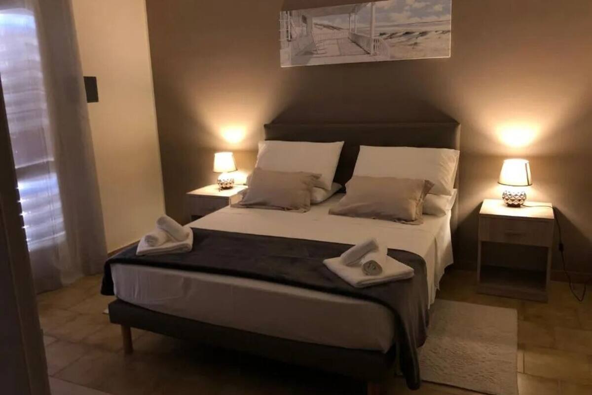 A neatly made bed with white linens and two pillows, a bedside table with a lamp, and a painting above the bed.