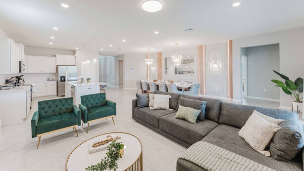 A spacious living area with a grey sofa, green armchairs, and a round coffee table. A kitchen with white cabinets and a dining area with a chandelier are visible in the background. - Stunning 8BR Pool Gameroom Minutes to Disney (Clermont)