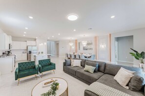 A spacious living area with a grey sofa, green armchairs, and a round coffee table. A kitchen with white cabinets and a dining area with a chandelier are visible in the background. - Stunning 8BR Pool Gameroom Minutes to Disney (Clermont)