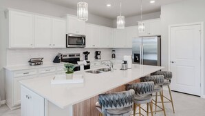 A modern kitchen with white cabinetry, stainless steel appliances, a large island with bar stools, and pendant lighting. - Stunning 8BR Pool Gameroom Minutes to Disney (Clermont)