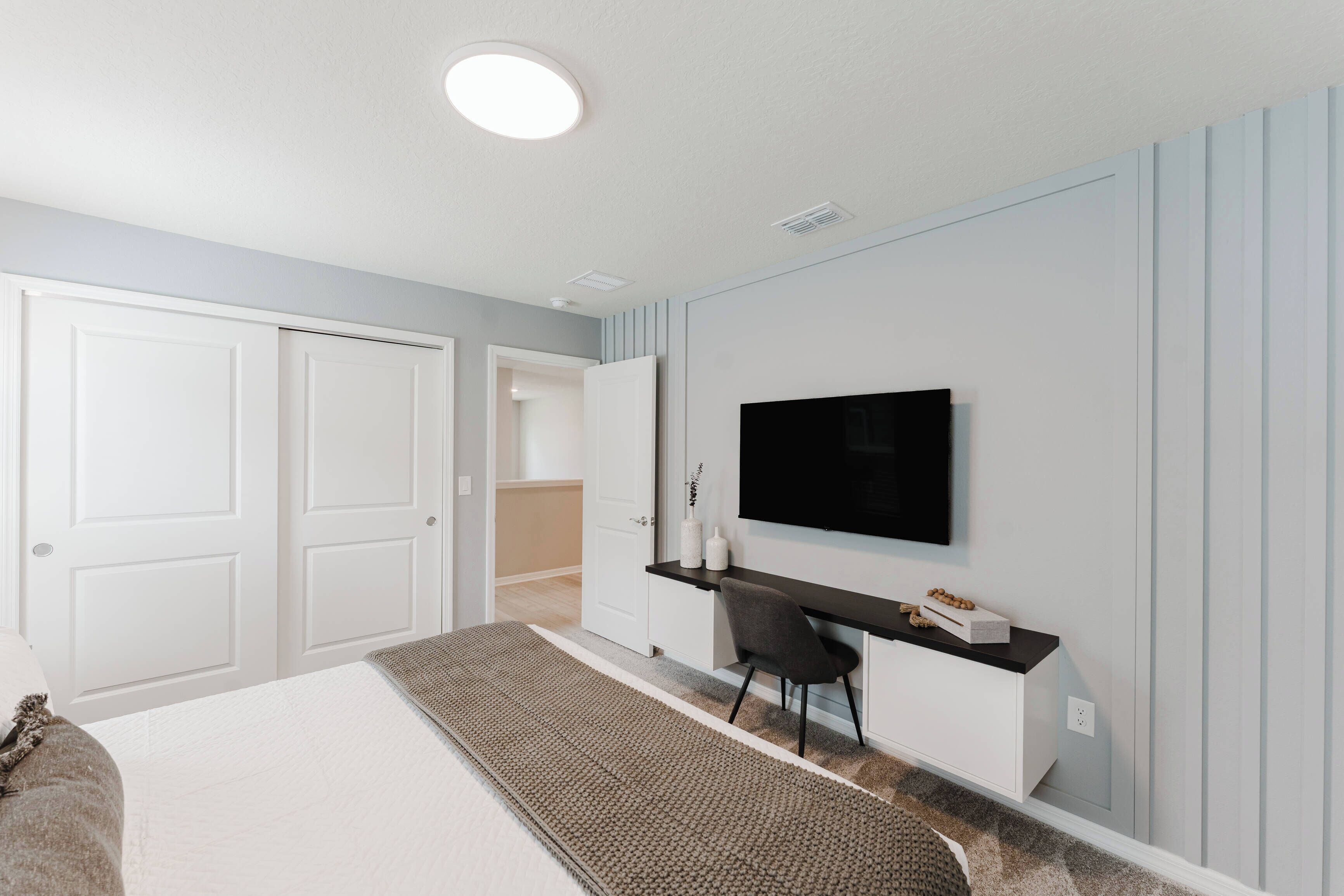 Un dormitorio con cama, escritorio, silla, televisor montado en la pared y una puerta.