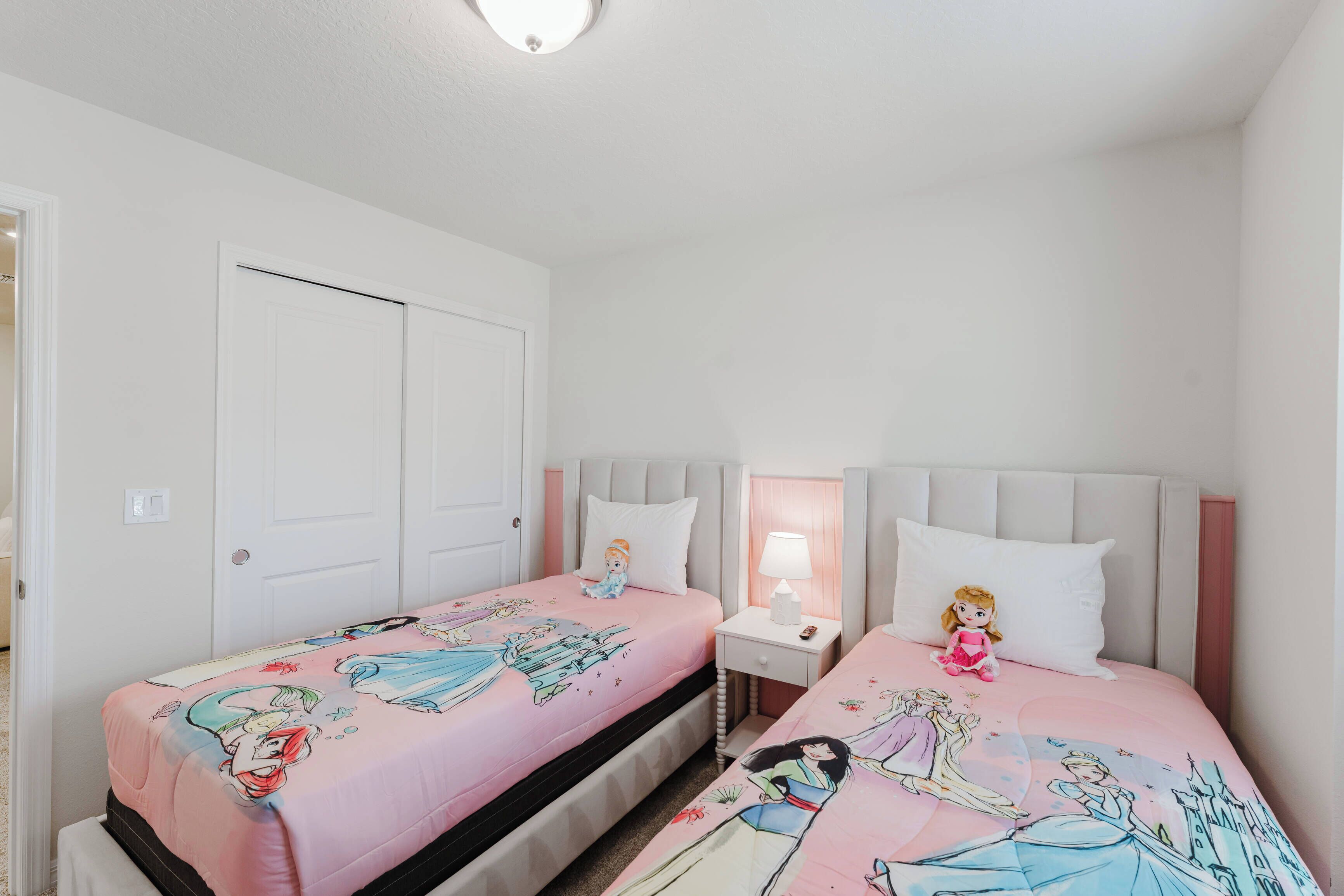 A bedroom with two beds, each with a pink bedspread featuring cartoon characters. There is a nightstand with a lamp between the beds.