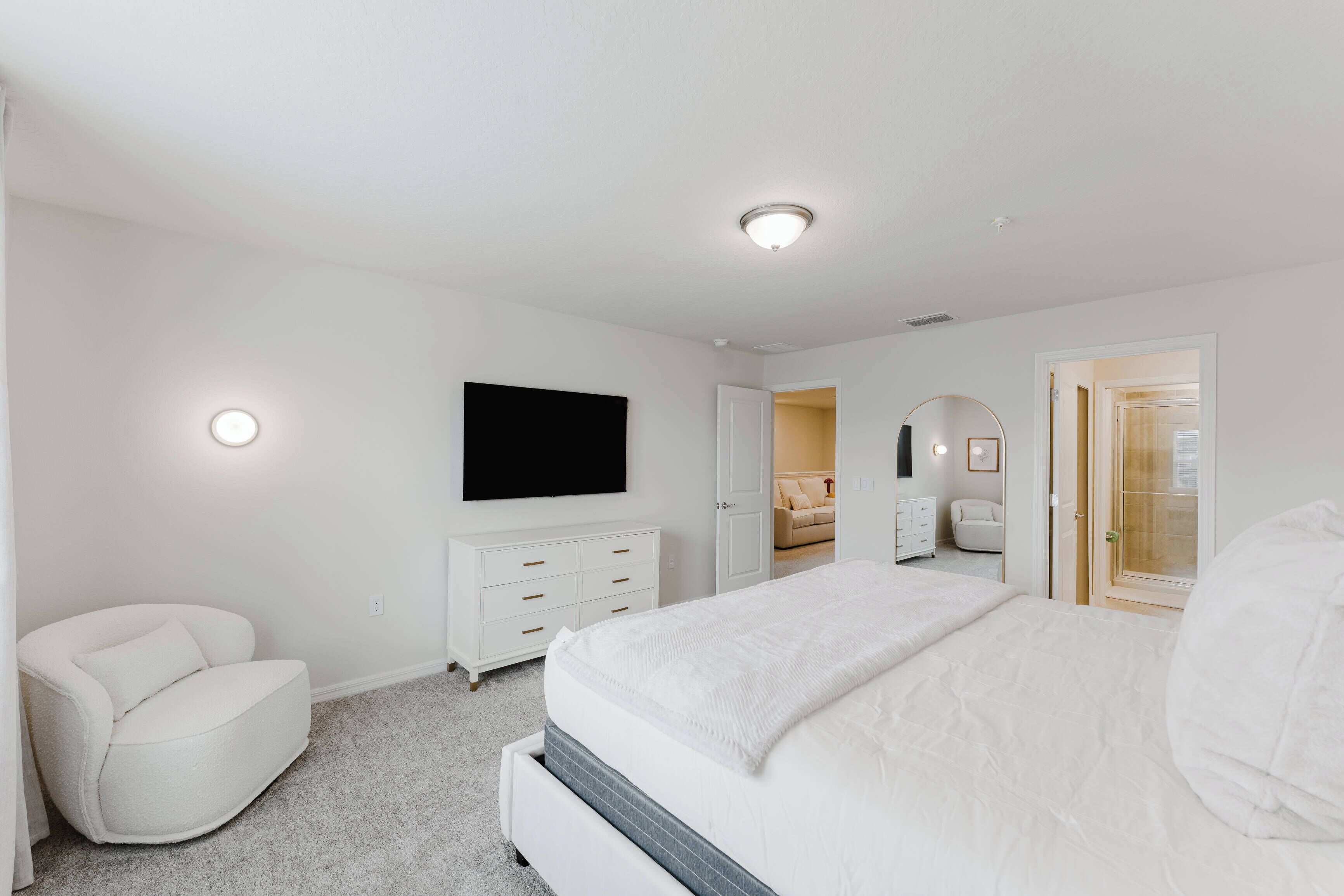 Un dormitorio moderno con una cama grande, un sillón blanco, un aparador y un televisor montado en la pared.