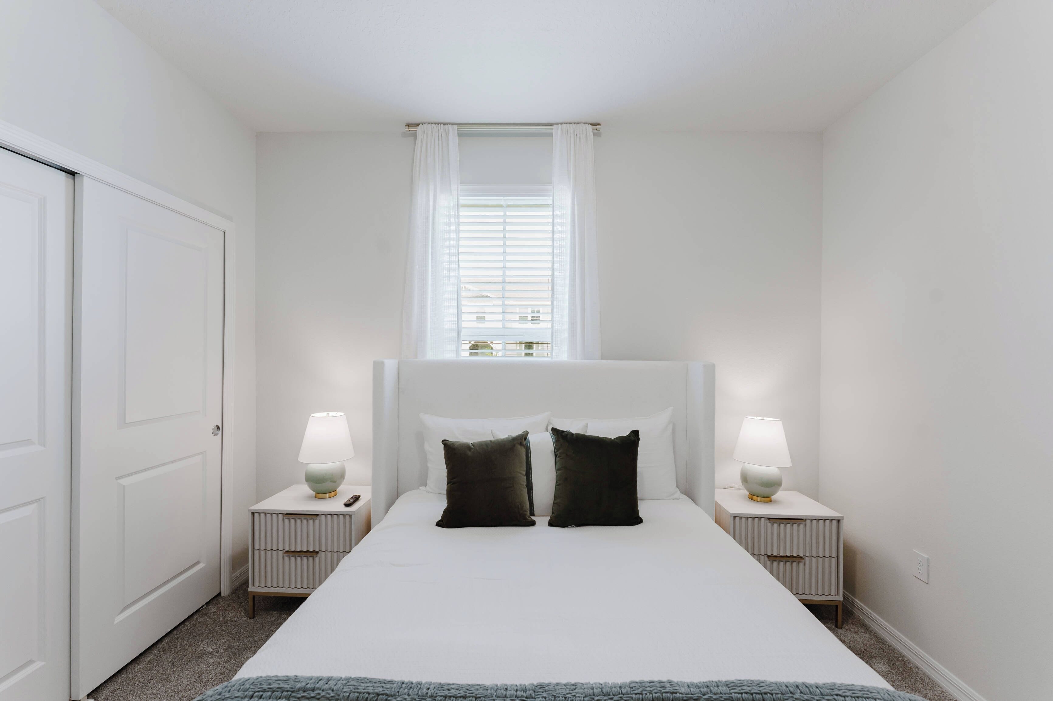 A bedroom with a large bed, two bedside tables with lamps, a window with blinds, and a door.