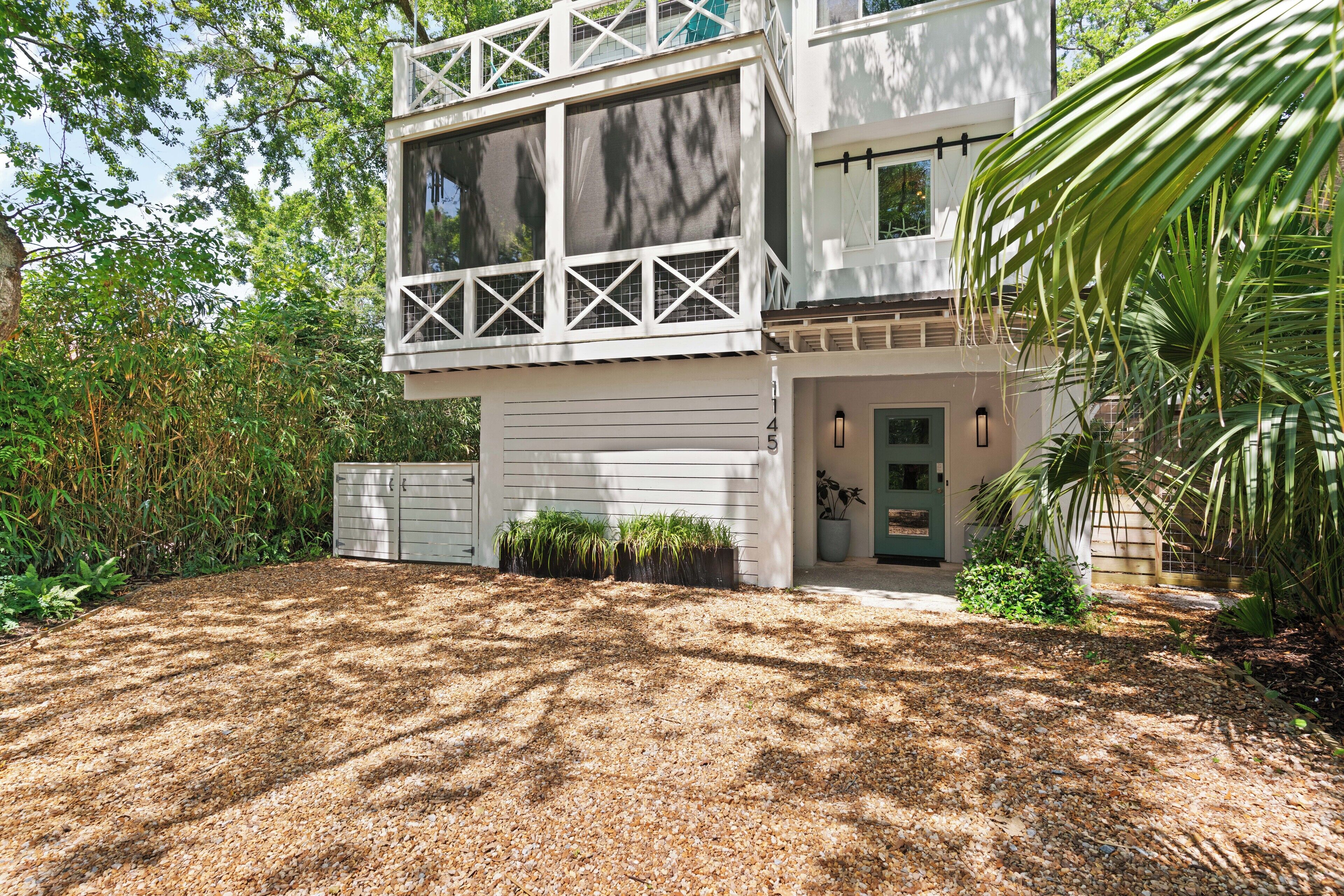 A modern two-story house with a large garage, surrounded by dense vegetation and a gravel driveway.