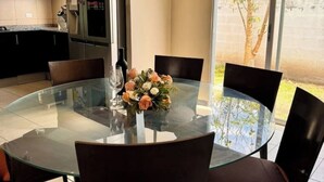 A round glass table with chairs, a vase of flowers, and a kitchen in the background.