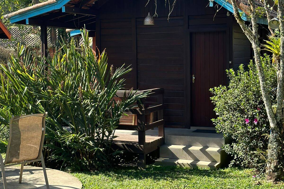 Una cabaña rústica de madera con techo inclinado, un porche con banca y un pequeño jardín con diversas plantas.