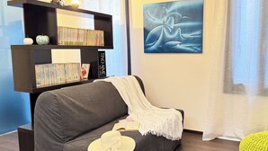 Ein Wohnzimmer mit grauem Sofa, einer Bücherwand, einem gerahmten abstrakten Gemälde und einem gelben Hocker.