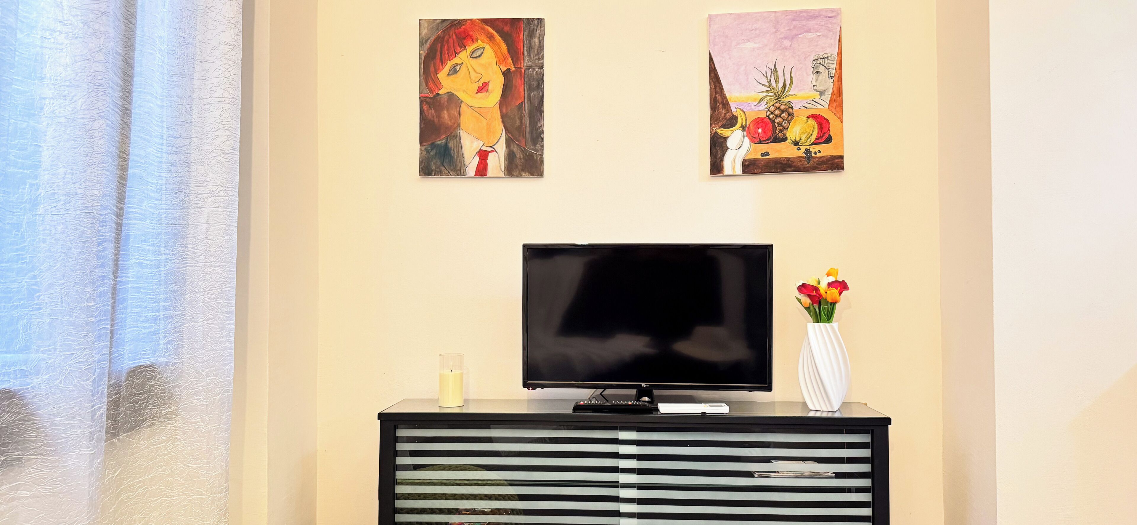 Une pièce avec un téléviseur posé sur un meuble noir, un vase blanc contenant des fleurs et deux tableaux accrochés au mur.