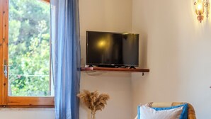 Una habitación con una silla de madera, un televisor montado en la pared, una ventana con cortinas y un jarrón decorativo con flores secas.