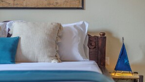 Una cama con cabecera de madera, un almohadón azul y un almohadón blanco. Junto a la cama hay una pequeña mesa con un paraguas azul.