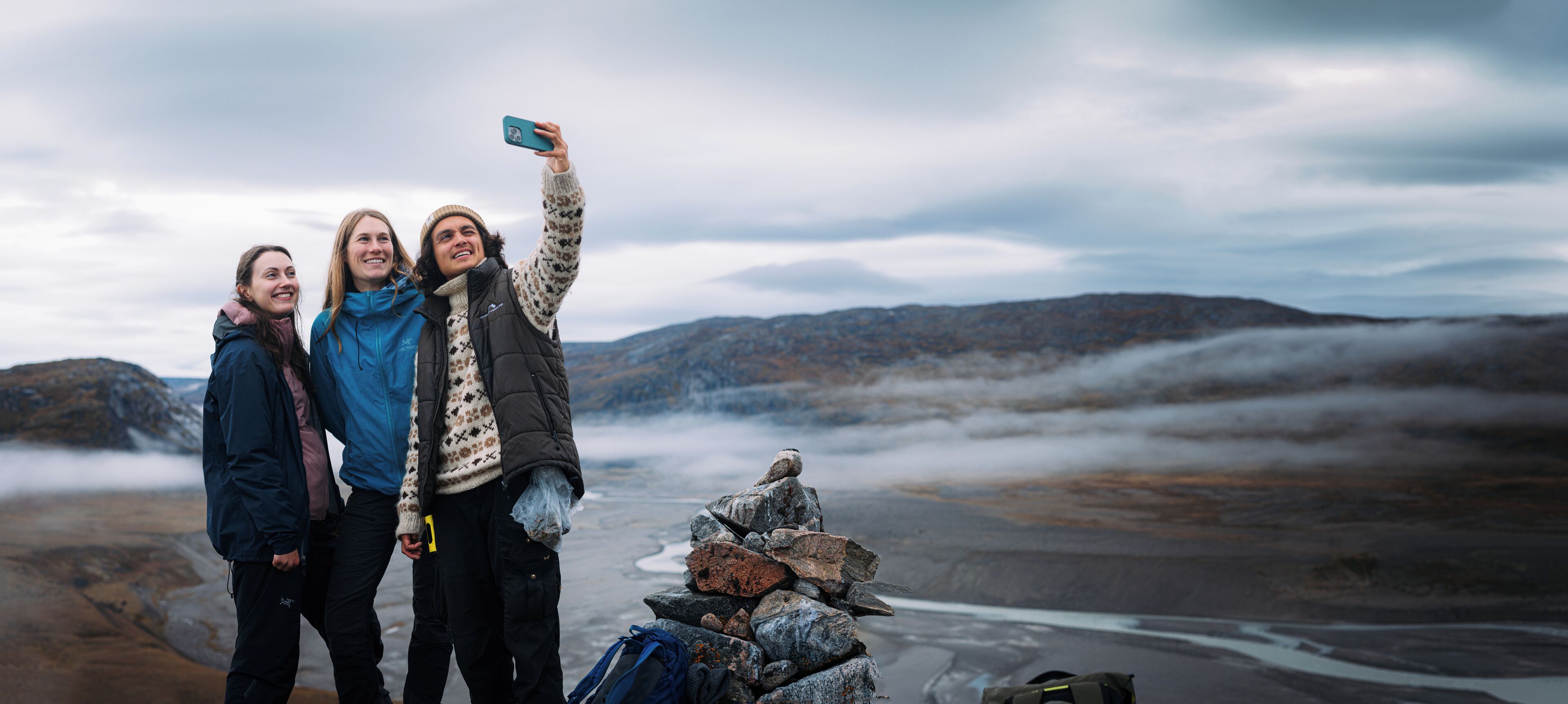 Trois personnes prennent un autoportrait devant un paysage rocheux, avec des montagnes en arrière-plan.