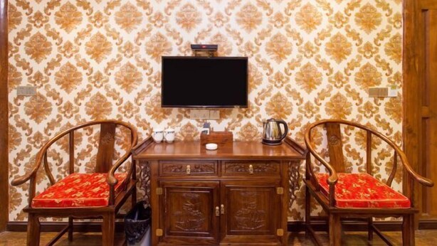 Ein Zimmer mit einem Holzschrank, zwei Stühlen mit roten Kissen, einem an der Wand montierten Fernseher und Tapete mit Muster.