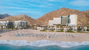 A beach resort with multiple hotel buildings, a sandy beach with sun loungers and umbrellas, and a mountainous backdrop.