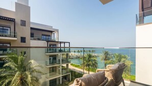 A balcony with a view of the ocean, palm trees, and a modern building with multiple floors.