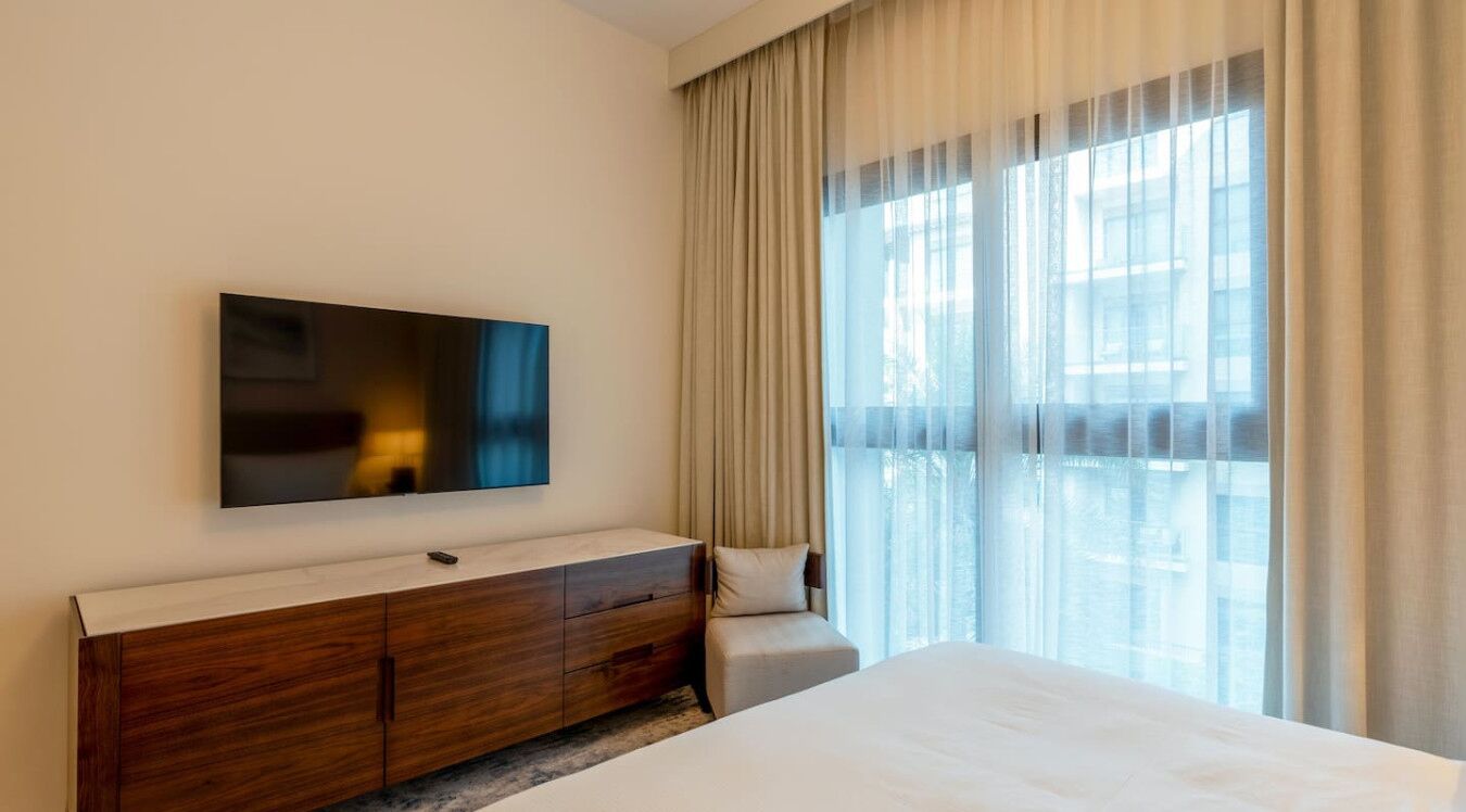 Une chambre d’hôtel avec un lit, une commode en bois, un téléviseur à écran plat fixé au mur et une grande fenêtre ornée de rideaux légers.