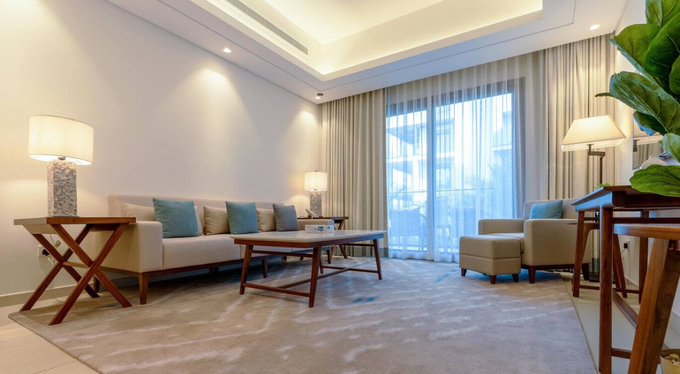 A modern living room with a sofa, coffee table, and armchairs. There are two lamps and a large plant.