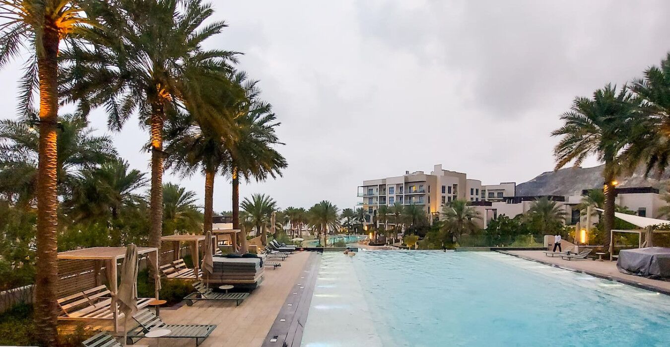 A pool area with lounge chairs and palm trees, a building with balconies in the background, and a clear sky.